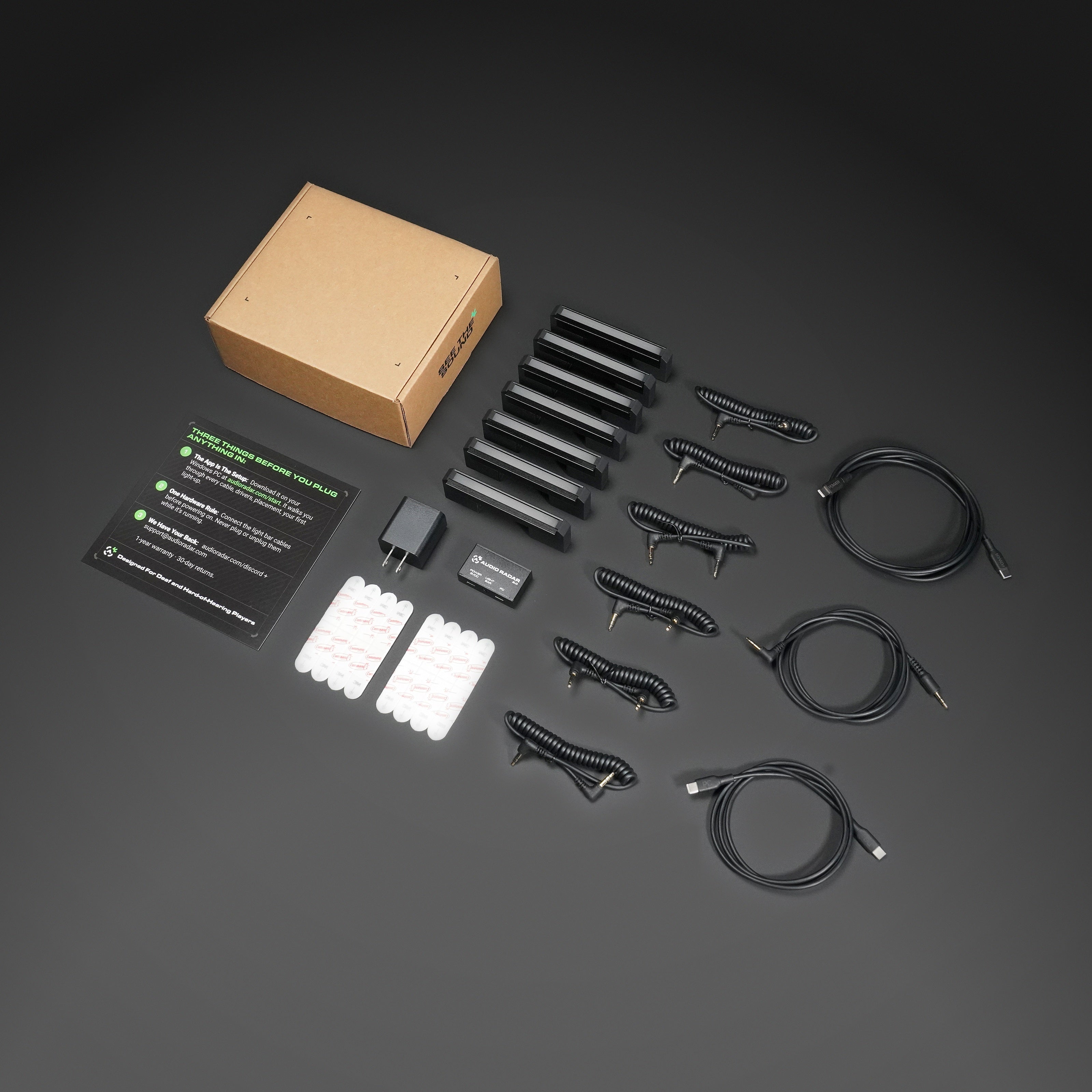 A complete kit of black USB-C to USB-C cables, power adapters, white adhesive pads, a brown cardboard box, and an instruction card, arranged neatly on a dark gray background.